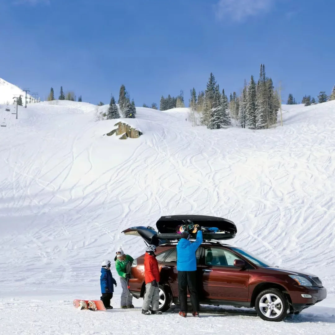 Family preparing winter gear next to a red SUV with a Thule Convoy XT Alpine cargo box in a snowy mountain setting.