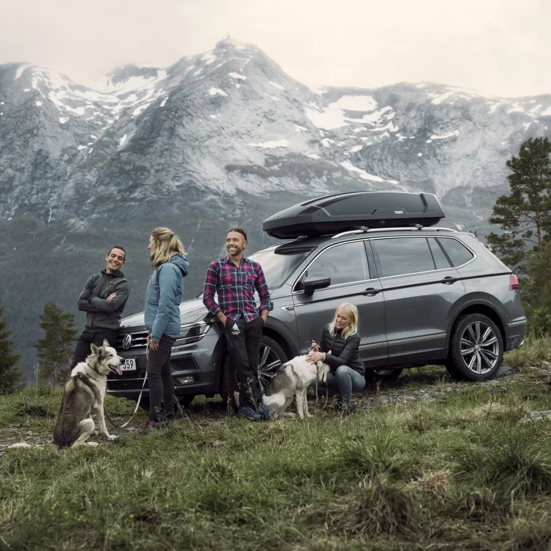 A family with dogs gathered near a parked SUV equipped with the Thule Force XT Sport box in a mountainous area.