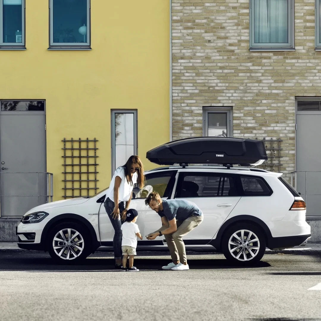 Family unloading luggage from an SUV with a Thule Force XT Sport box parked in front of colorful buildings.