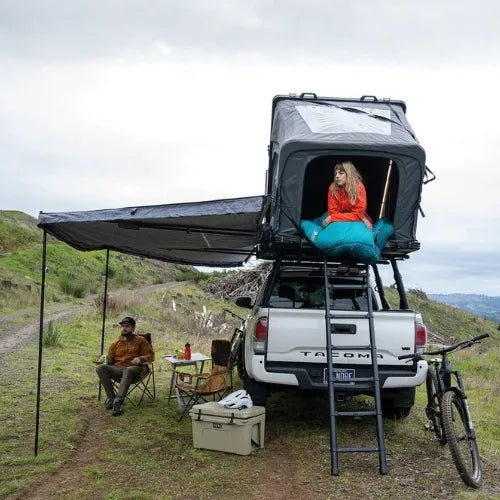 Camper truck with open roof rack and canopy, people sitting outside on a trail.