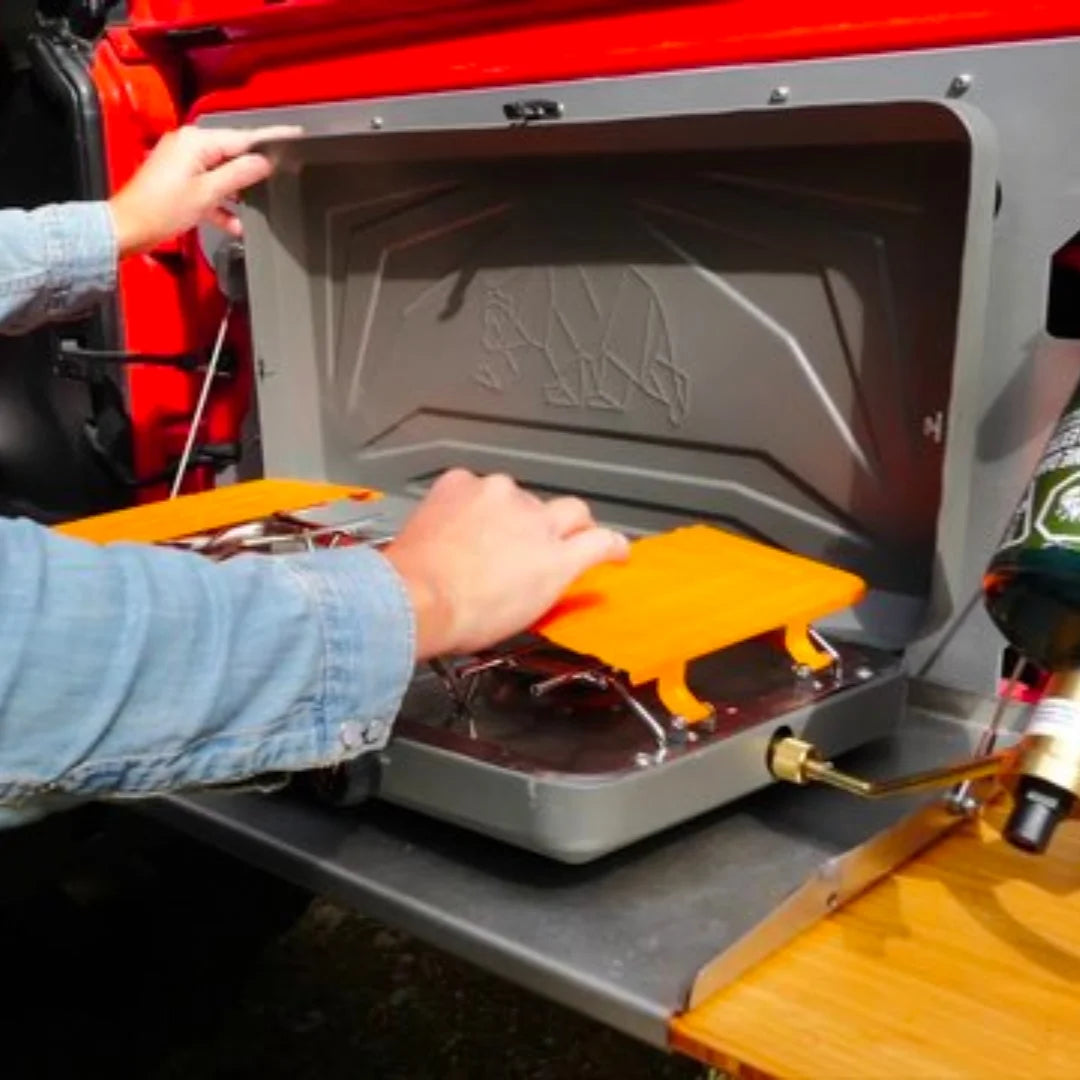 Person adjusting controls on Kuma 2-burner stove while cooking outdoors on a wooden table