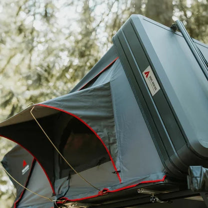 Gray rooftop tent with red accents attached to a vehicle, set against a blurred natural background.