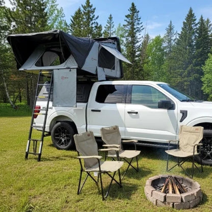 White truck with a rooftop tent in a forest setting with chairs and a fire pit.