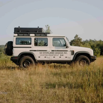 White SUV with folded rooftop tent and 'GO Overland Canada Inc.' branding parked in a grassy field.