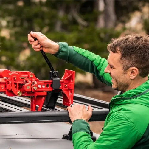 Man in green jacket using a red car jack on a vehicle outdoors.