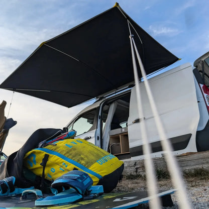 Van with open door, black awning extended, and camping gear on the ground.
