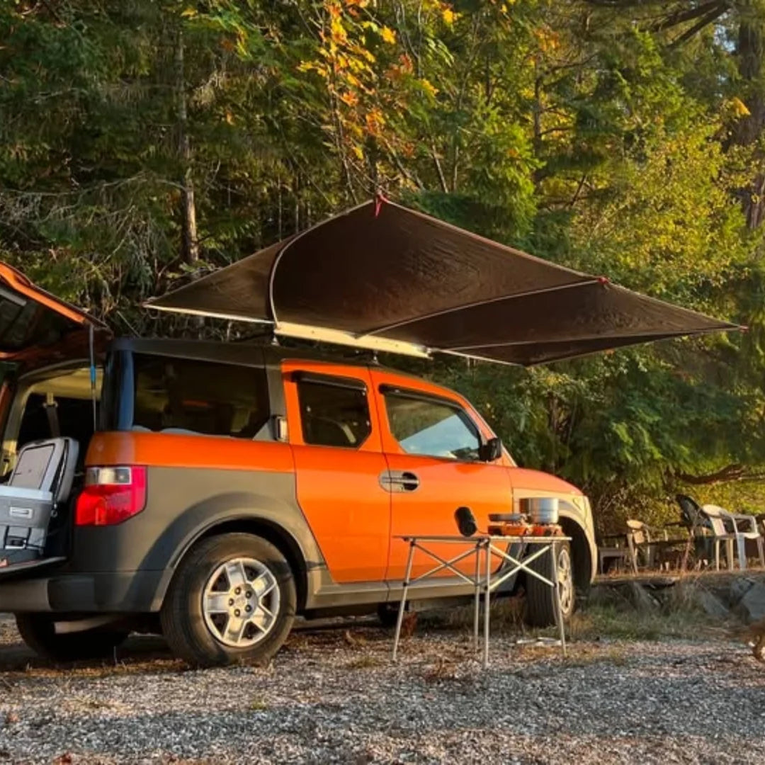 Orange truck with a shady boy rooftop awning deployed and parked in a forest setting with a sunset