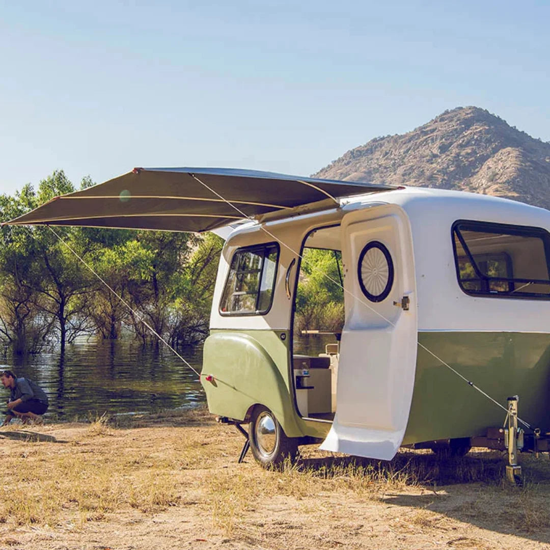 Vintage-style camper van with an extended awning parked near a body of water with mountains in the background.