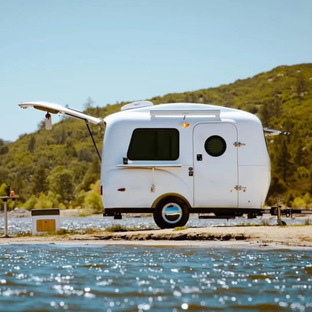 Small white camper trailer parked by a body of water with mountains in the background and folded shady boy awning attached to it