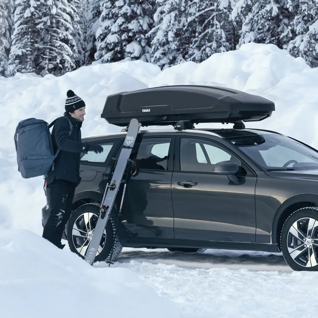  Person in snow gear loading gear into a Thule Force XT Sport box mounted on a vehicle in a snowy forest.