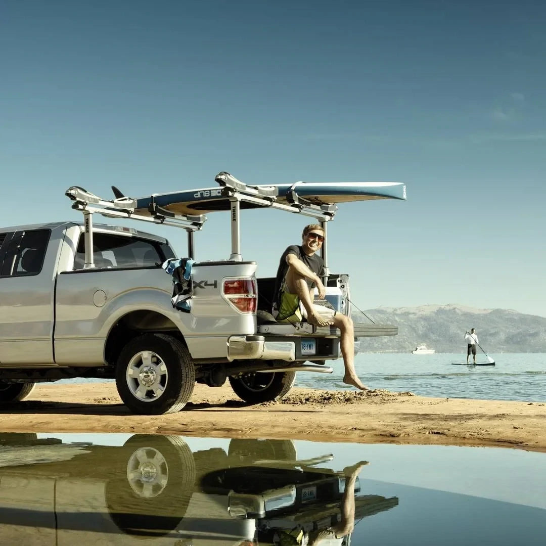 Thule Xsporter Pro setup on a white pickup truck parked by water, loaded with surfboards, with person looking happy