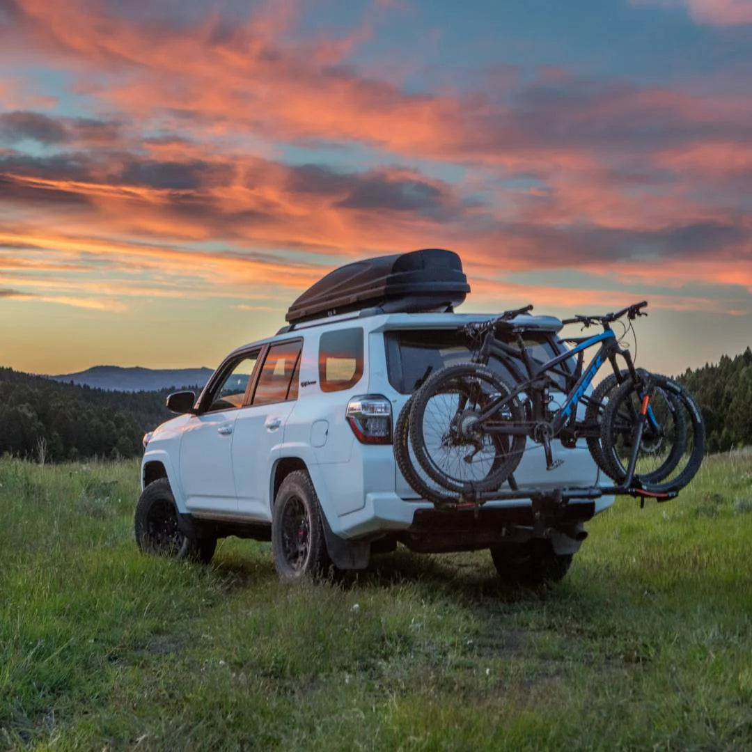 Scenic photo of SUV with Kuat rack and bikes near mountain sunset.

