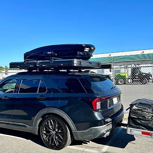 Black SUV with rooftop tent and cargo box on crossbars, pulling hitch trailer for overlanding