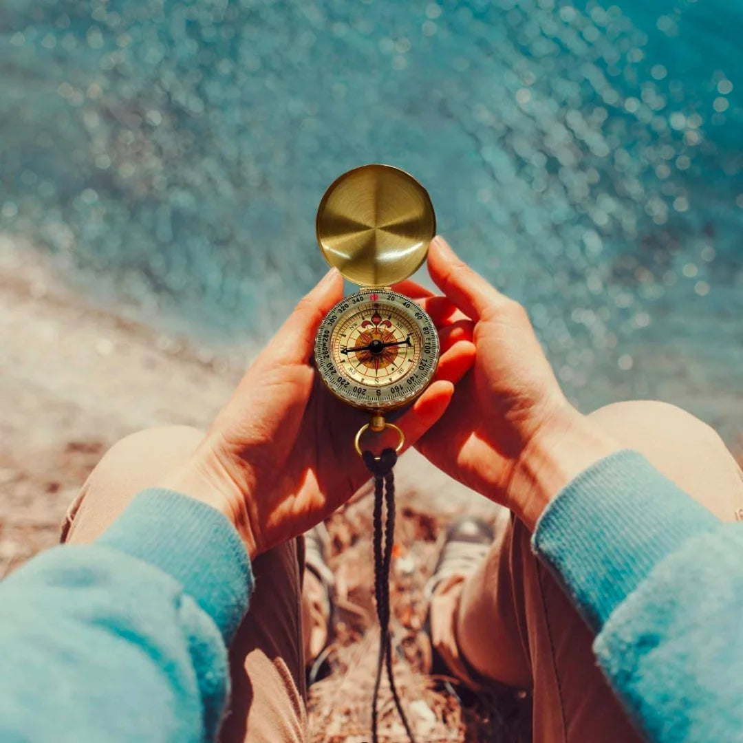 Person holding AO brass tactical compass outdoors with ocean background.