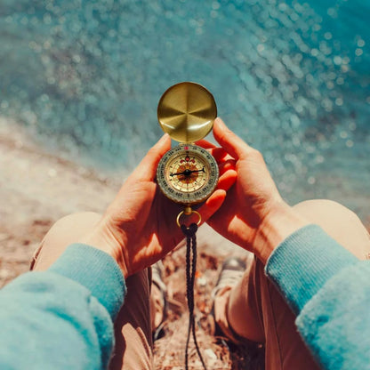 Person holding AO brass tactical compass outdoors with ocean background.