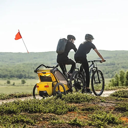 Two cyclists towing Thule Chariot Sport Single yellow trailer on off-road trail