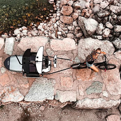 Overhead view of Thule Reacha Sport bike trailer connected to bicycle carrying kayak on rocky shore path, emphasizing adventure use