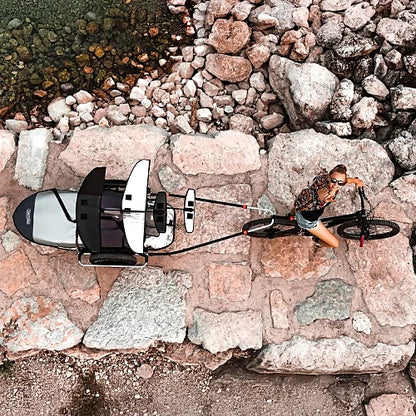 Overhead view of Thule Reacha Sport bike trailer connected to bicycle carrying kayak on rocky shore path, emphasizing adventure use