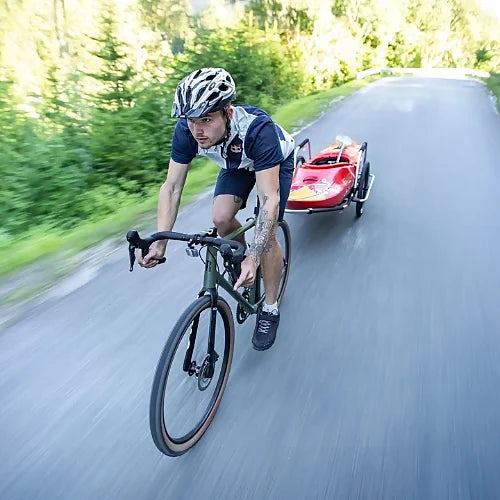 Cyclist riding with Thule Reacha Sport trailer carrying red kayak on paved road, showcasing aerodynamic design and durability