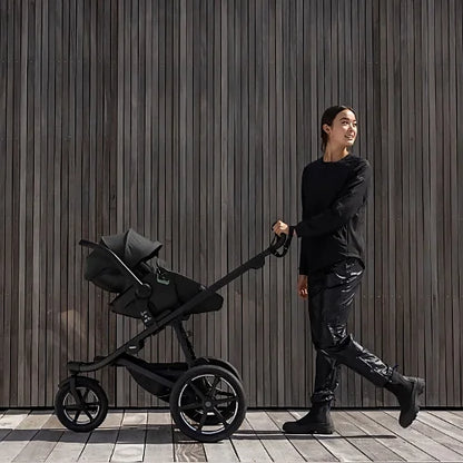 Parent pushing Thule Urban Glide 3 black all-terrain stroller along wooden wall background