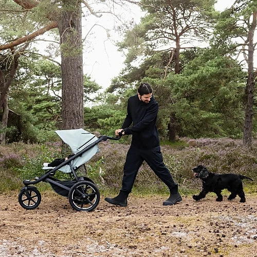 Parent pushing Thule Urban Glide 3 stroller on forest trail while walking a dog
