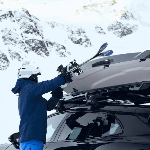 Person loading skis into Thule Vector Alpine roof box showing dual-side opening and convenient gear access in snowy mountains