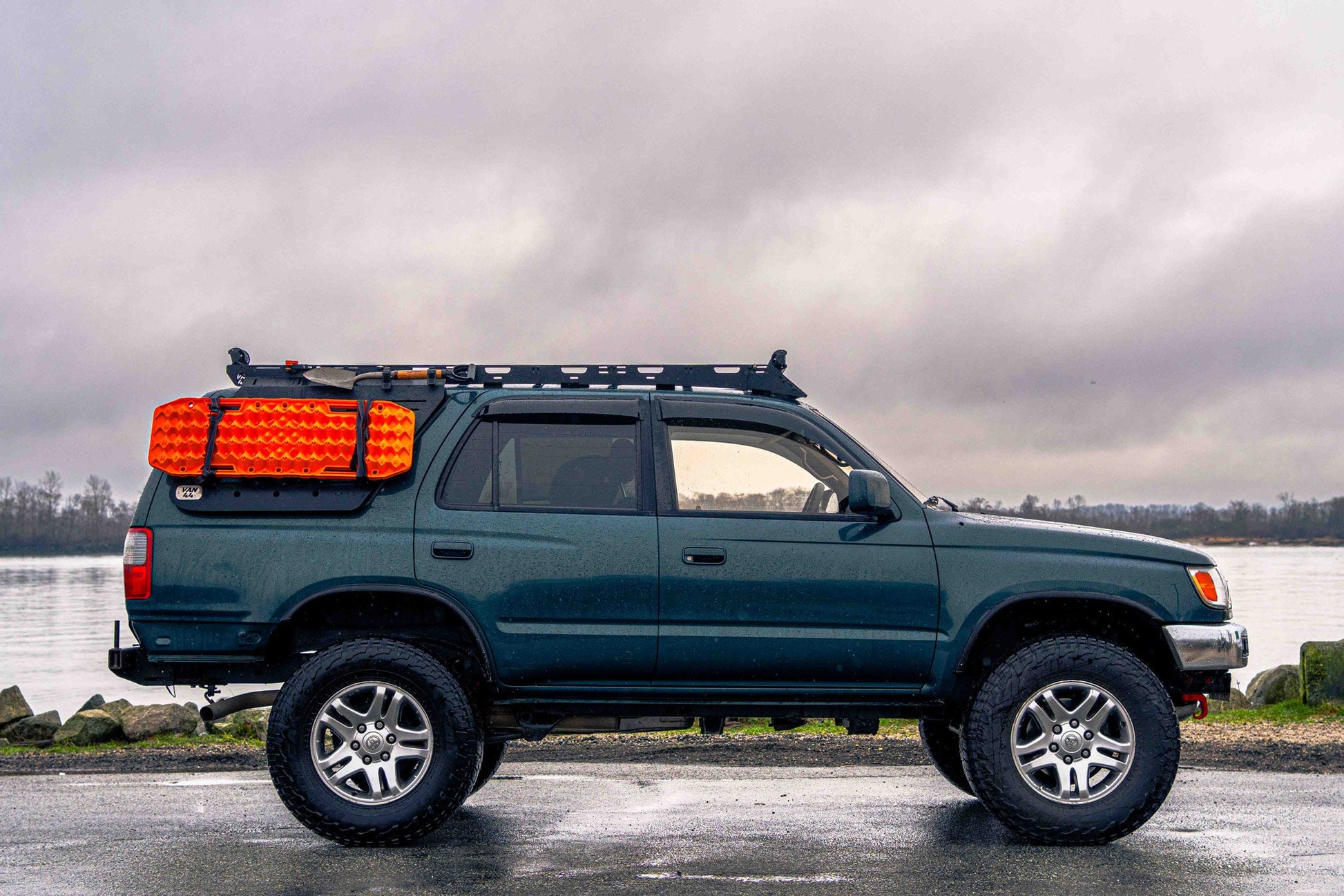 Blue SUV with van44 platform roof rack and orange traction boards parked by a body of water on a cloudy day.