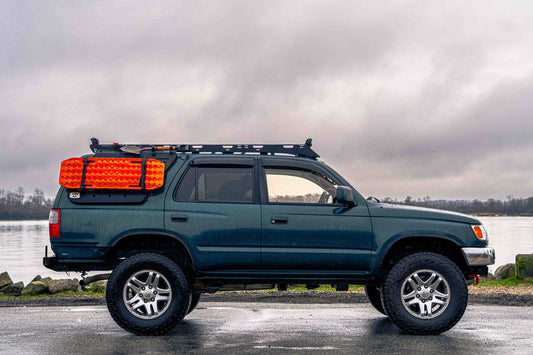 Blue SUV with van44 platform roof rack and orange traction boards parked by a body of water on a cloudy day.
