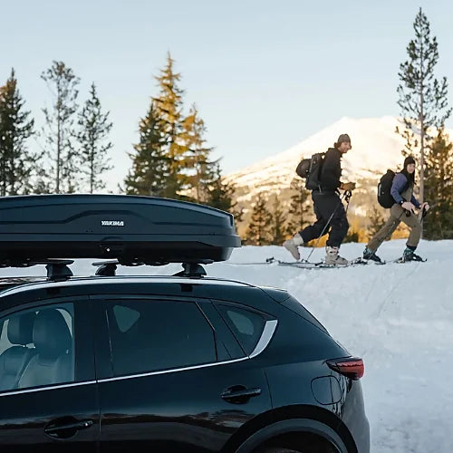Yakima SkyBox NX Skinny roof box mounted on SUV with skiers walking in snowy mountain setting